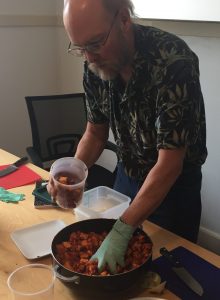 Bruce is holding his container with one hand and reaching into the batch of freshly tossed radishes with the other hand during the kimchi workshop. 