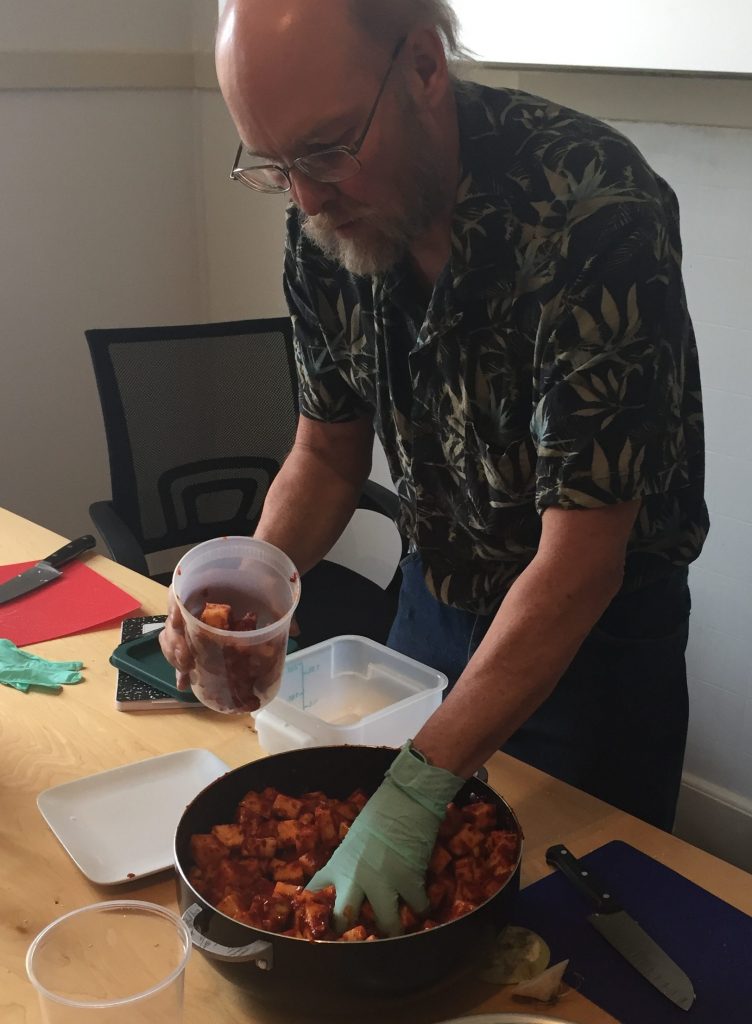 Bruce is holding his container with one hand and reaching into the batch of freshly tossed radishes with the other hand.