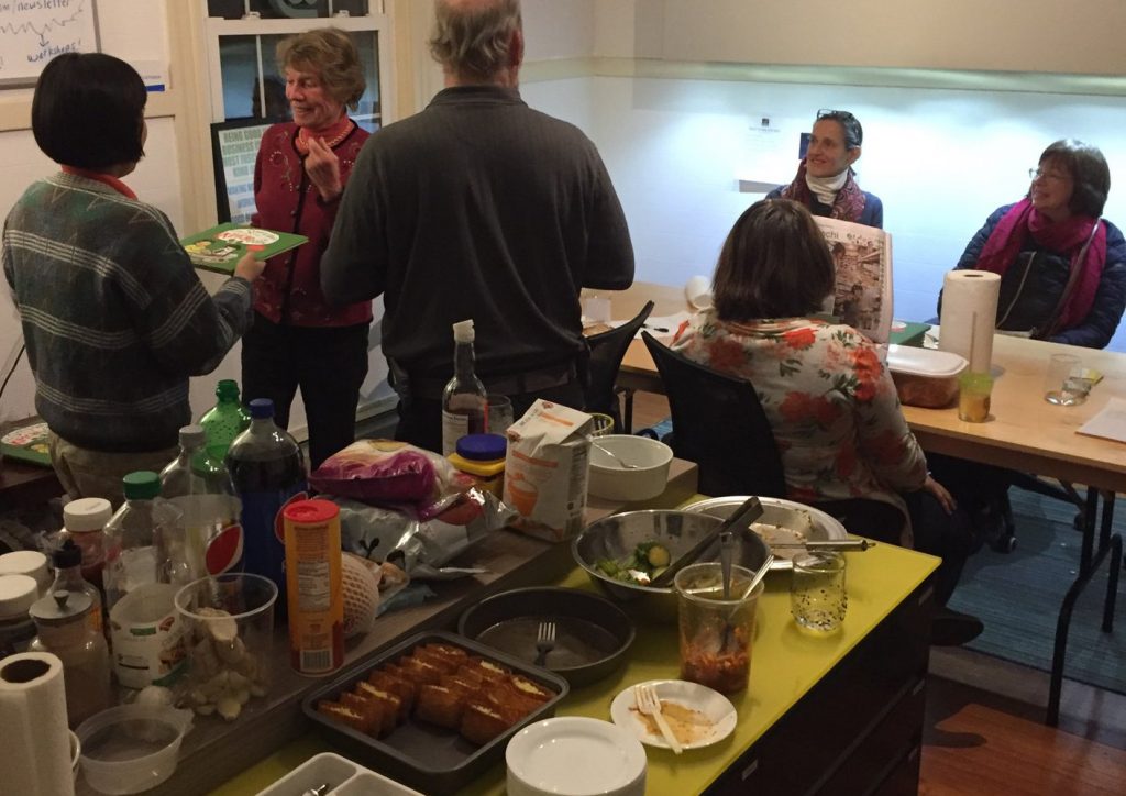 Counter is full of condiments and party food; Jill is handing a book to Scotty looking pleased; Doug is standing with his back toward the camera; Nicole is reading a newspaper at the table; Betsy and Joan are sitting at the table, looking towards Jill and Scotty.