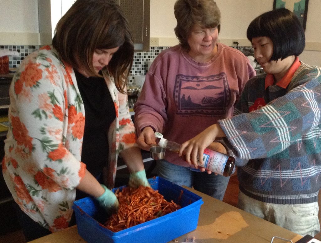 Some kimchi workshop action: Nicole has her gloved hands in the paste, Kelley is holding a measuring cup out over the paste bucket, and Jill is pouring fish sauce into Kelley's cup.