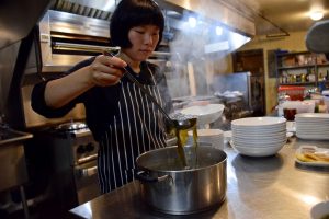 Jill is in a metal-clad kitchen, about to ladel seaweed soup into a bowl. This photo was used as a part of the article about Little Sunny in the news.